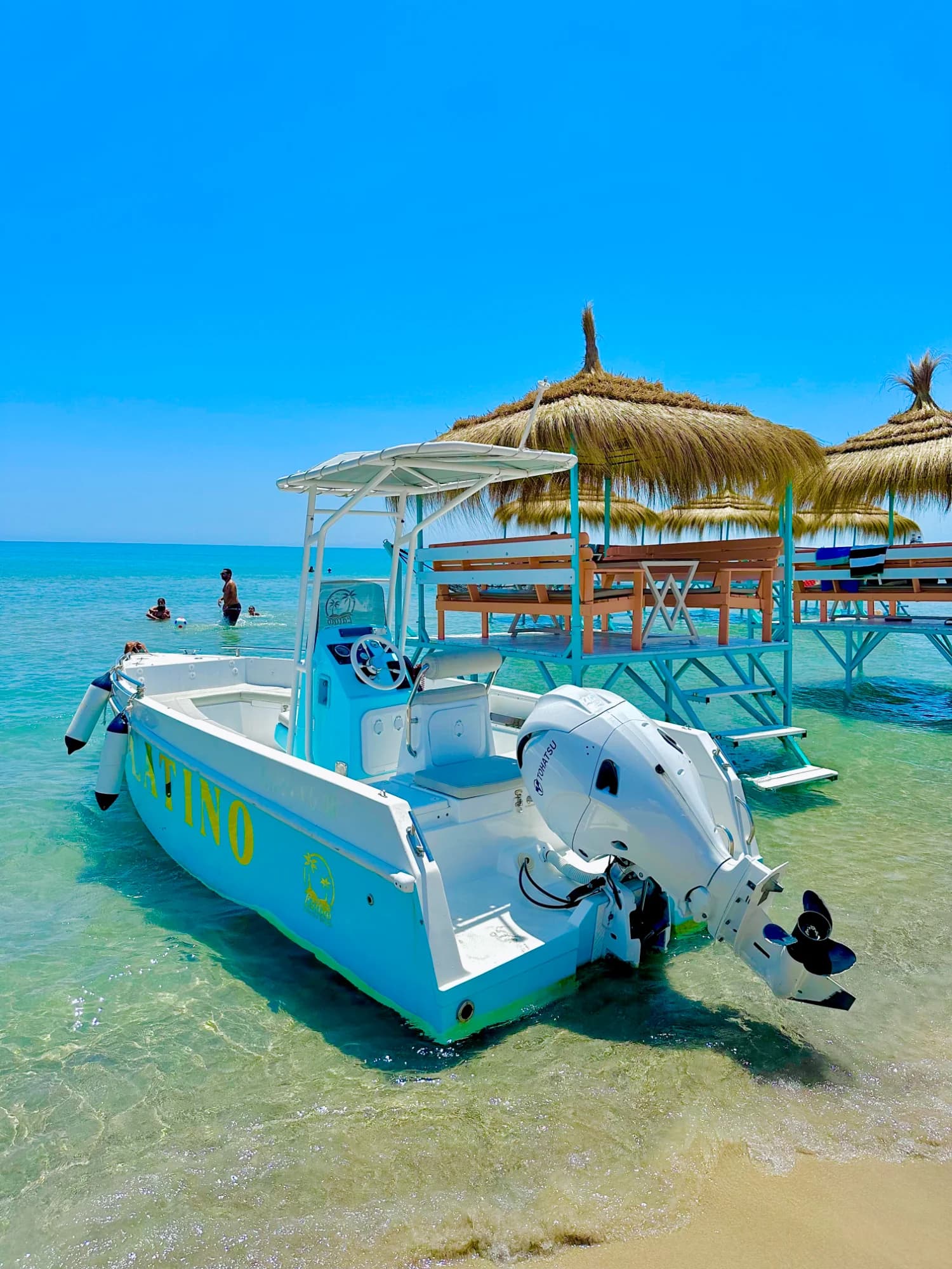 un bateau-moteur blanc et bleu avec le mot 'LATINO' écrit en jaune, amarré dans l'eau cristalline près de parasols en paille sur des chaises longues, sous un ciel bleu vif à la plage de latino coucou beach.