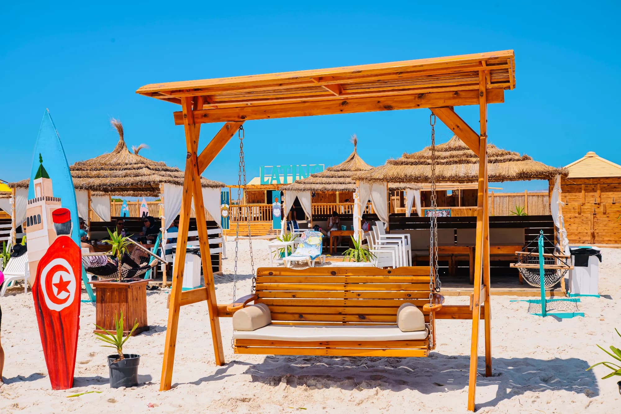 Une balançoire en bois avec coussins beiges sur une plage de sable, entourée de cabanes avec toits en paille et un panneau de surf rouge et bleu orné du drapeau tunisien, sous un ciel bleu.