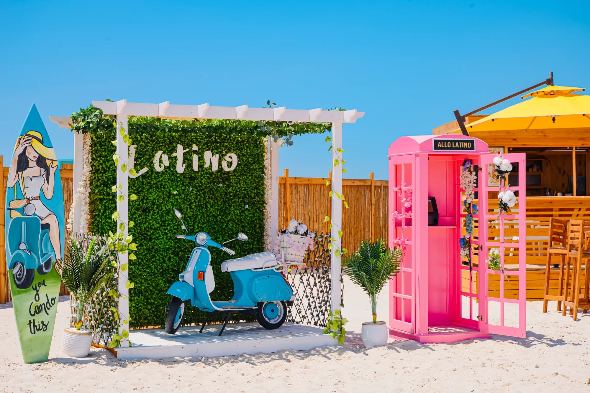 scooter vespa de couleur bleue et boîte téléphonique rose vif situés sur une plage décorée avec une arche verdure et une planche de surf latine
